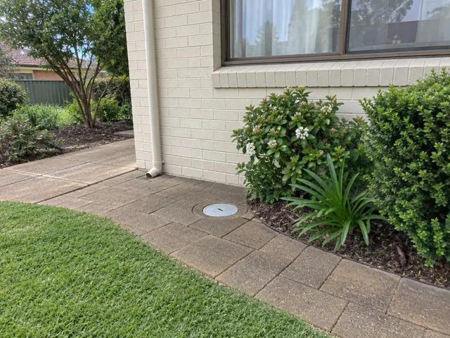 Capped access point beside paved path and garden beds next to a house wall in Kingswood