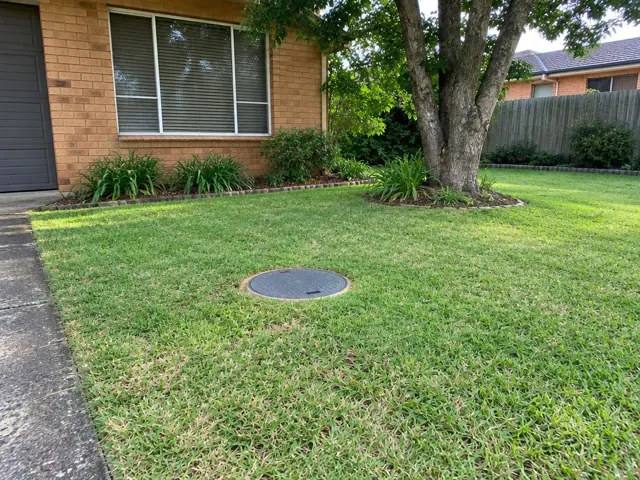 Round sewer access cover set flush in a suburban lawn beside a brick house in St Marys
